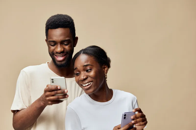 Young African couple laughing together while using smartphones against a neutral beige backdrop, capturing joy and connection through technology