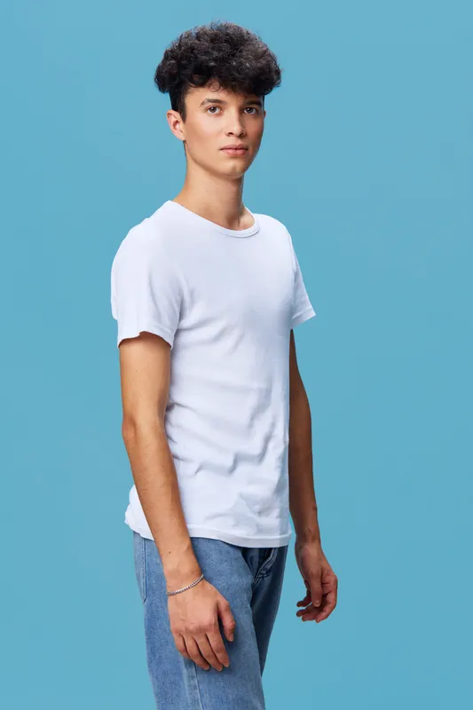 young man wearing white t-shirt and blue jeans stands against plain blue background with calm expression and casual posture, showcasing simple fashion style