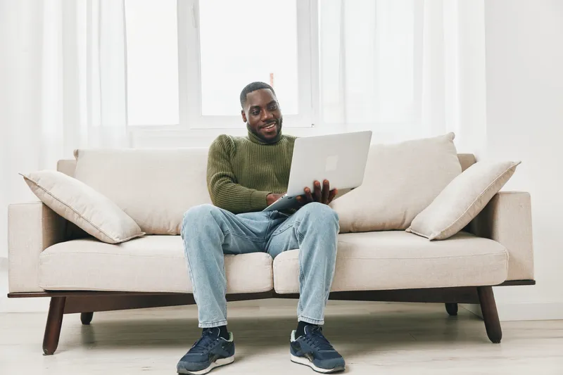 Happy young Black man using laptop on a cozy sofa in a bright, modern living room, showcasing a relaxed and productive atmosphere during home office hours