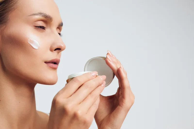 Young woman applying white face cream on cheek with eyes closed, holding a round jar. Concept of skincare, beauty routine, moisturizing, and self-care with soft lighting.