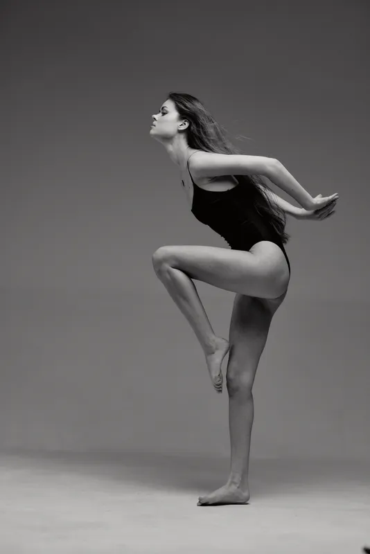 Black and white image of a graceful woman dancer posing on one leg, showcasing elegance and balance in a minimalist studio setting.