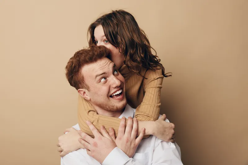 Happy young couple in casual clothes embraces and smiles against beige background. Woman hugs man from behind showing affection and joy. Concept of love, relationship, and togetherness.