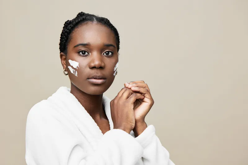 Young Black woman applying skincare in a cozy robe, showcasing healthy skincare routine and natural beauty with soft neutral background