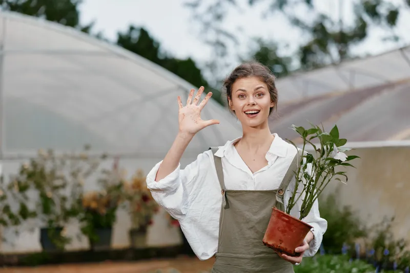 Young cheerful woman in overalls holding a plant pot, celebrating gardening success in a greenhouse with vibrant greenery