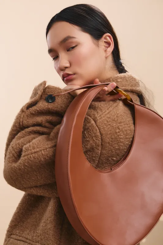 Asian woman models a chic mood with a warm coat and oversized bag, posing with a soft gaze in a studio, conveying style, elegance, and calm confidence.