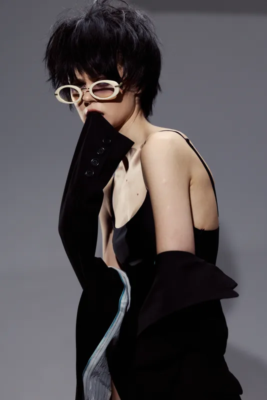 Stylish woman wearing sunglasses and black outfit posing with hand near mouth in studio lighting