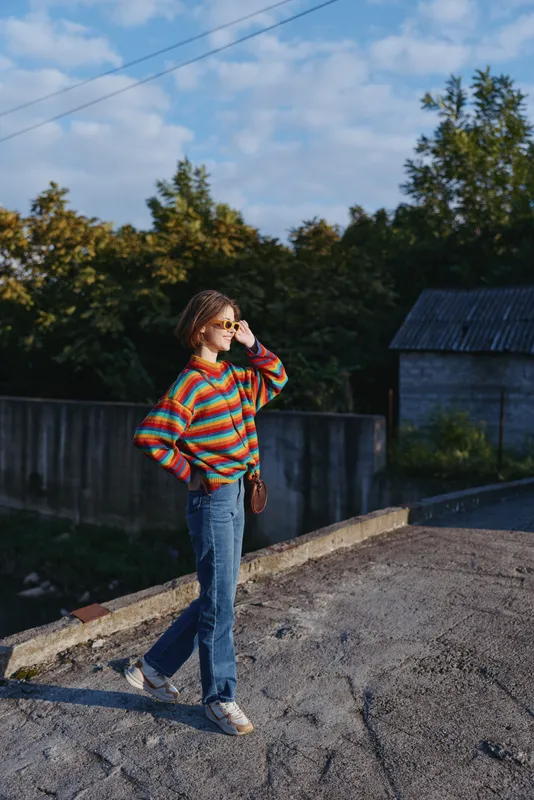 Young woman in her 20s wearing a colorful striped sweater and denim jeans, walking and smiling while adjusting sunglasses, crossbody bag and casual outfit in a suburban outdoor lifestyle fashion
