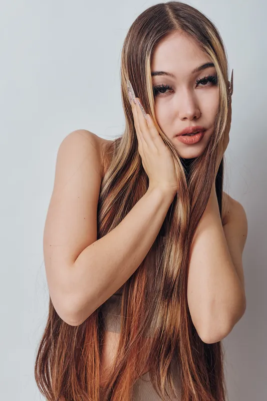 Woman with long brown hair and braids posing with hands near face on gray background. Female model with smooth skin and natural makeup looking at camera in studio portrait.