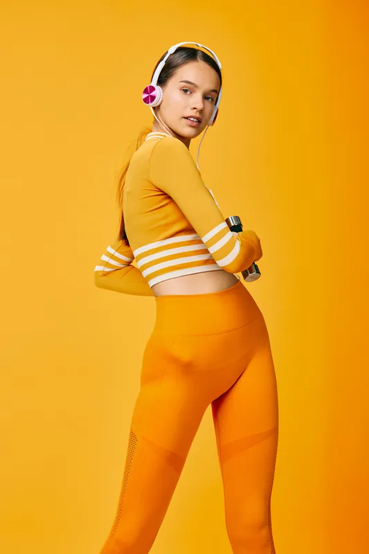 Athletic young woman in orange activewear and headphones poses confidently against a vibrant orange background, showcasing fitness, sporty style, training readiness and modern wellness.