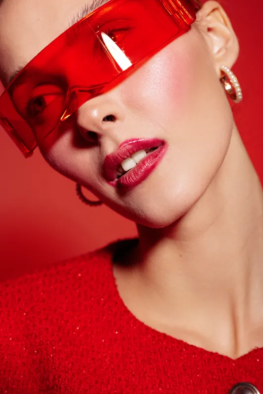 Close-up of a fashionable woman wearing red translucent visor and red sweater. She has glossy lips and hoop earrings, posing against a red background. Concept of modern style and bold fashion.
