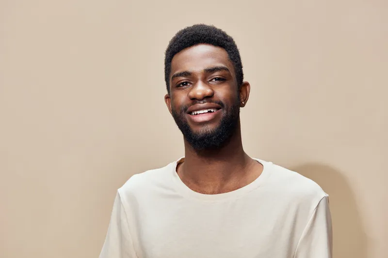 Young Black man smiling against a neutral beige background, wearing a casual light t shirt, conveying joy and confidence in a contemporary style