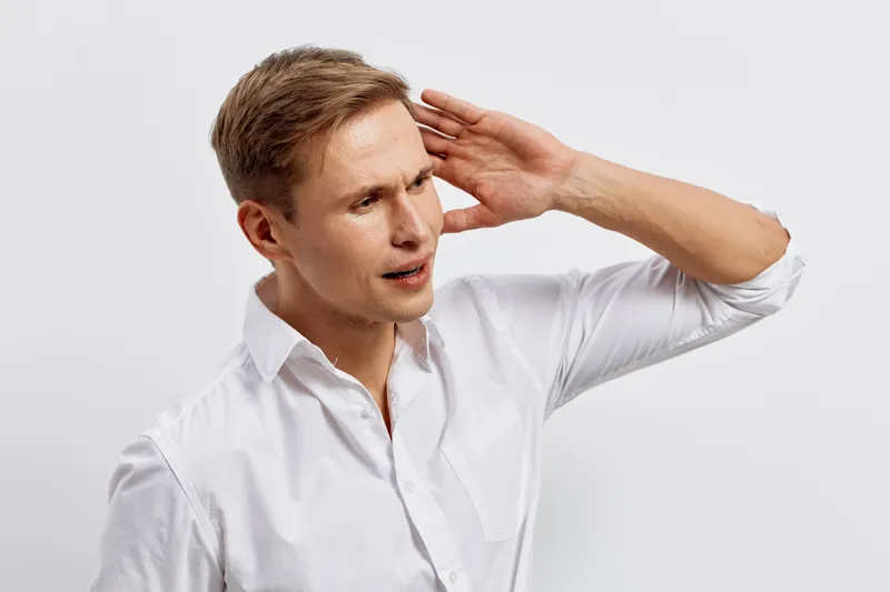 Professional portrait of a young man with light brown hair wearing a white shirt, holding his hand to his head in a gesture of confusion or hearing difficulty, isolated on a plain white background. Pe