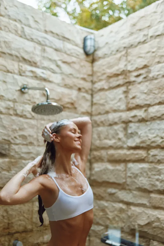 woman showering outdoors in sunlight with soap foam, enjoying refreshing water under stone wall, natural skincare and wellness moment in warm atmosphere