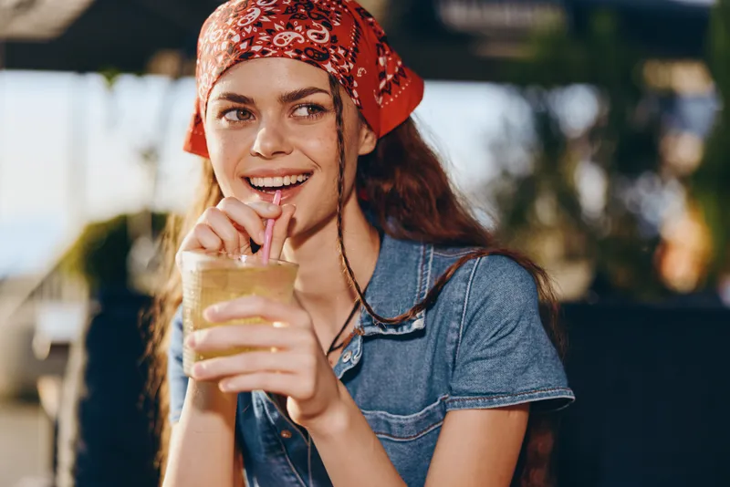 Lifestyle woman in modern boho-western denim street style sipping a drink with a playful smile, featuring warm film color and casual outdoor summer vibes.
