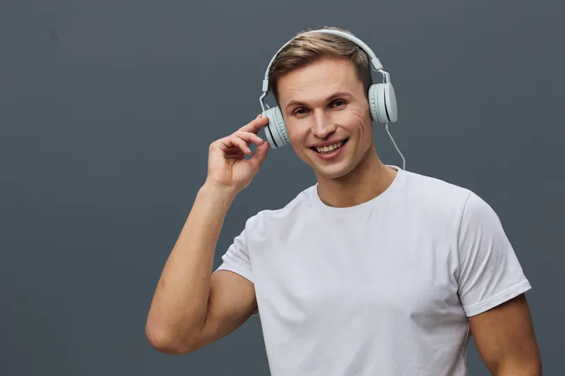Smiling young man with headphones, casual white t-shirt, enjoying music, studio shot on gray background, cheerful expression, lifestyle concept, modern technology.