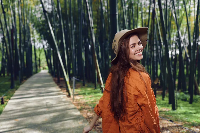 Woman smiling and walking on a paved path through a bamboo forest, wearing a wide hat and orange jacket, looking back over her shoulder during an outdoor nature stroll.