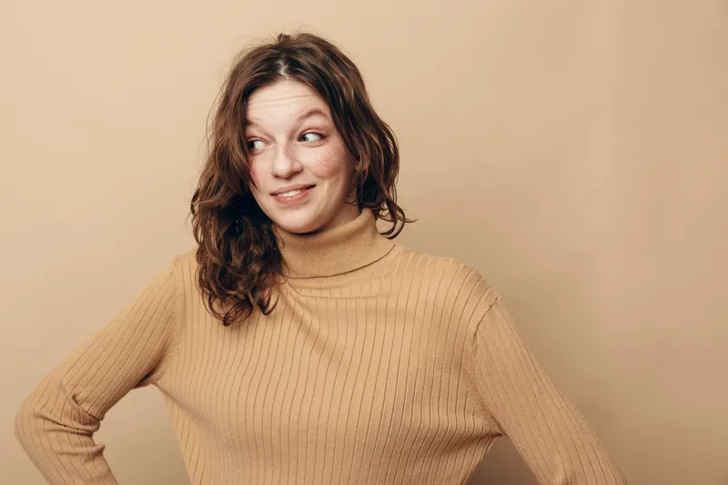 Young woman with curly hair wearing beige turtleneck sweater looks sideways with a playful smile. Neutral background and soft lighting create a warm, casual portrait concept.