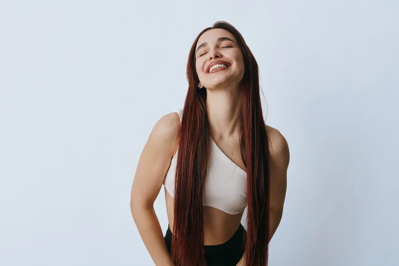 Young woman with long braided hair smiling with eyes closed wearing white crop top and black pants on light background. Happiness, beauty, casual style and confident pose.