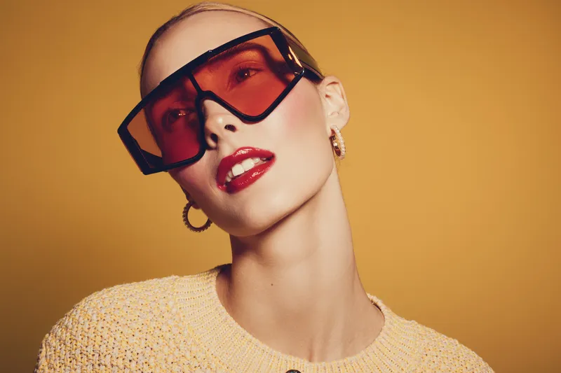 Stylish woman with red lipstick and large orange sunglasses poses against yellow background. Fashion portrait with bold makeup and trendy accessories in warm studio lighting.