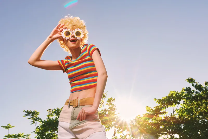 Happy young woman in a striped crop top wears flowered sunglasses, enjoying sunny outdoors, retro film color vibe, carefree lifestyle portrait against bright sky.