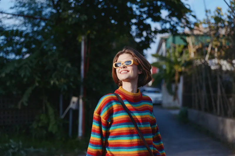 Woman in a colorful striped sweater smiling in sunlight, wearing sunglasses and a crossbody bag. Short hair, casual outfit on a suburban street for lifestyle travel photography.