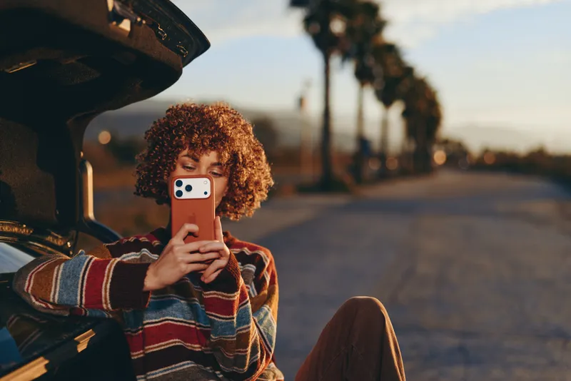Woman lifestyle selfie outdoors at sunset, holding smartphone up for selfie, curly hair and striped sweater, standing near open car door on quiet street, soft bokeh light