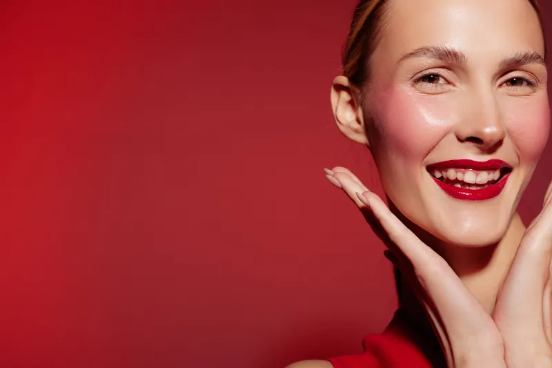 Smiling woman with red lipstick and blush poses with hands near face on red background. Close-up portrait with bright makeup and joyful expression. Concept of beauty and fashion.