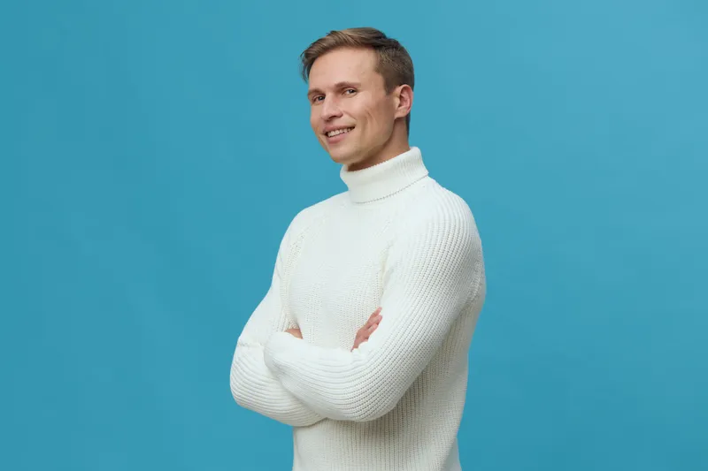 Smiling young man wearing cozy white turtleneck sweater posing against bright blue background studio portrait. Casual fashion lifestyle concept, cheerful expression, casual style, winter clothing, mal