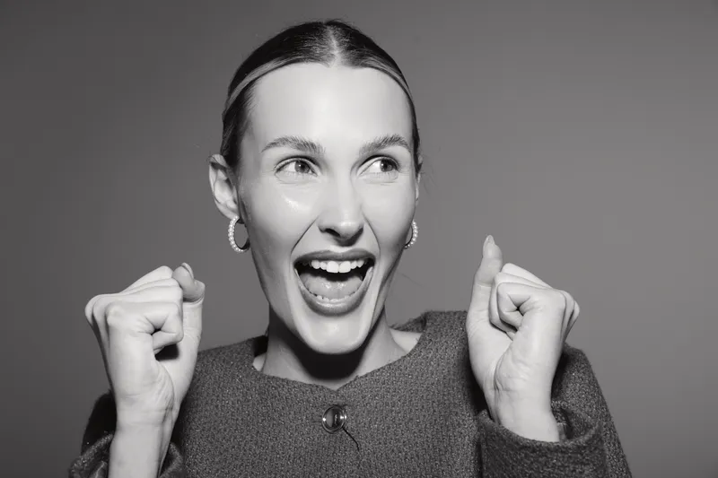 Caucasian woman with light skin expresses excitement and joy with wide open mouth and raised fists. Black and white studio portrait with emotional facial expression.