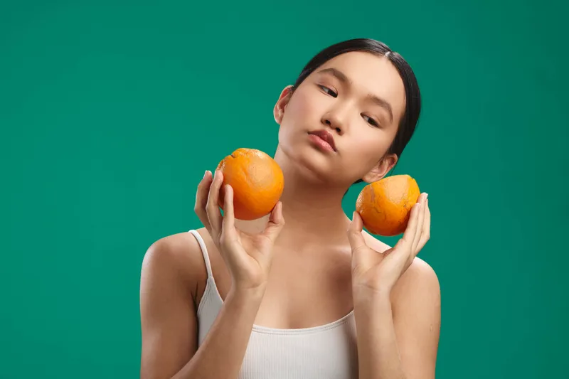 An asian woman stands against a vibrant backdrop, holding two oranges near her shoulders, expressing calm confidence and playful balance in a studio portrait.