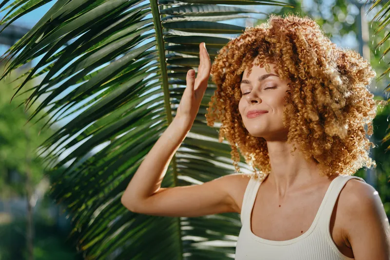 Young woman with curly hair enjoying sunlight outdoors near tropical palm leaves, wearing white tank top, expressing calmness and relaxation in nature.
