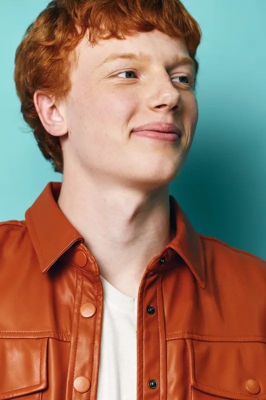 Young man with red hair wearing brown leather jacket and white shirt looking aside with slight smile on blue background. Casual style and relaxed expression with natural light.