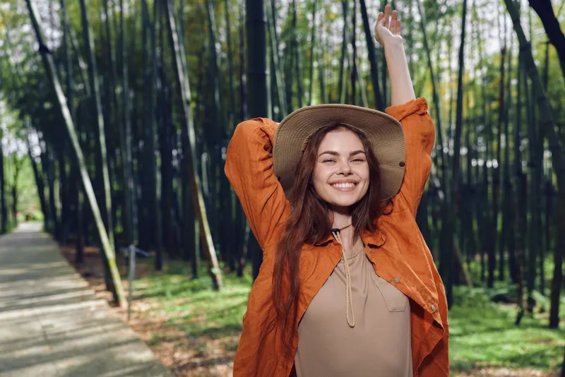 Woman smile hat forest nature happy outdoor portrait of a young woman in straw hat on a forest path, relaxed and carefree with orange jacket, arms raised in joyful weekend lifestyle moment.