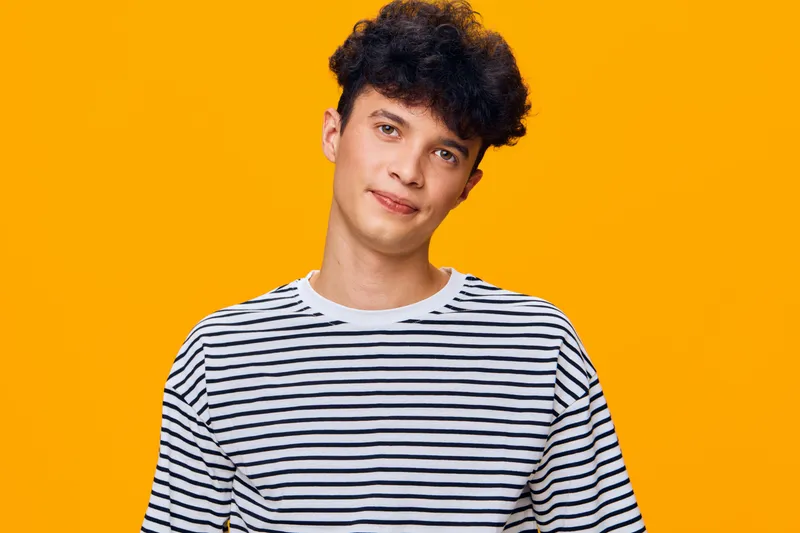 young man wearing striped shirt poses confidently against vibrant orange background, displaying casual style and relaxed expression in portrait photography.
