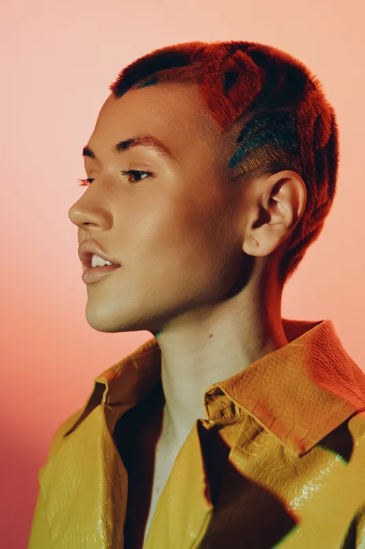 young woman with a short colorful hairstyle wearing a yellow jacket, posing in profile against a stylish warm background, modern fashion portrait
