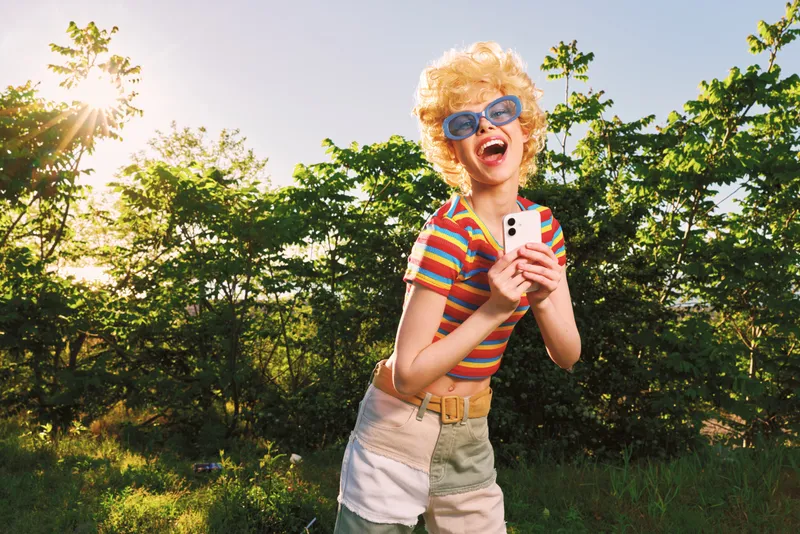 Young woman in a bright striped shirt and denim shorts laughs outdoors, holding a smartphone in a sunlit field with a retro film color palette and carefree energy.