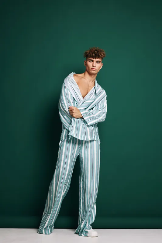 Young man standing in striped pajamas with open collar against green background, casual pose and relaxed expression in studio lighting for fashion or lifestyle concept.