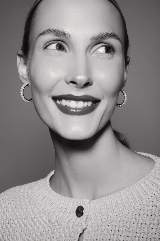 Close-up of a smiling young white woman with fair skin and sleek hair wearing hoop earrings and a knitted sweater in studio lighting.