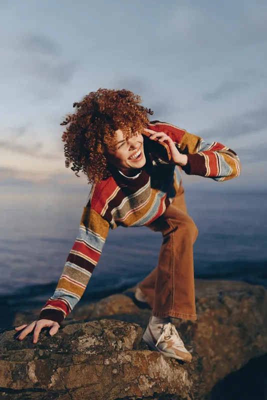 Smiling woman with curly hair wearing a striped sweater by the ocean rocks. Outdoor seaside lifestyle on a cloudy day, relaxed pose, soft natural light, warm mood for travel vacation.