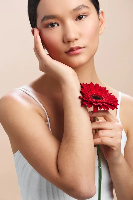 asian woman rests her cheek on her hand, holding a bright red flower against a soft studio backdrop, creating a calm, graceful portrait with gentle emotion and harmonic lighting.