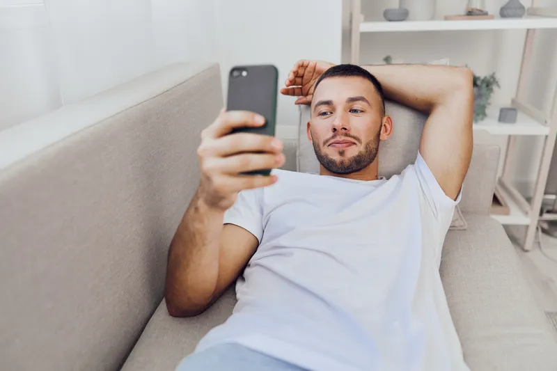 Relaxed man enjoying a moment of leisure while checking his smartphone, wearing casual clothing in a cozy living room setting. Vibrant colors and modern decor enhance the scene.