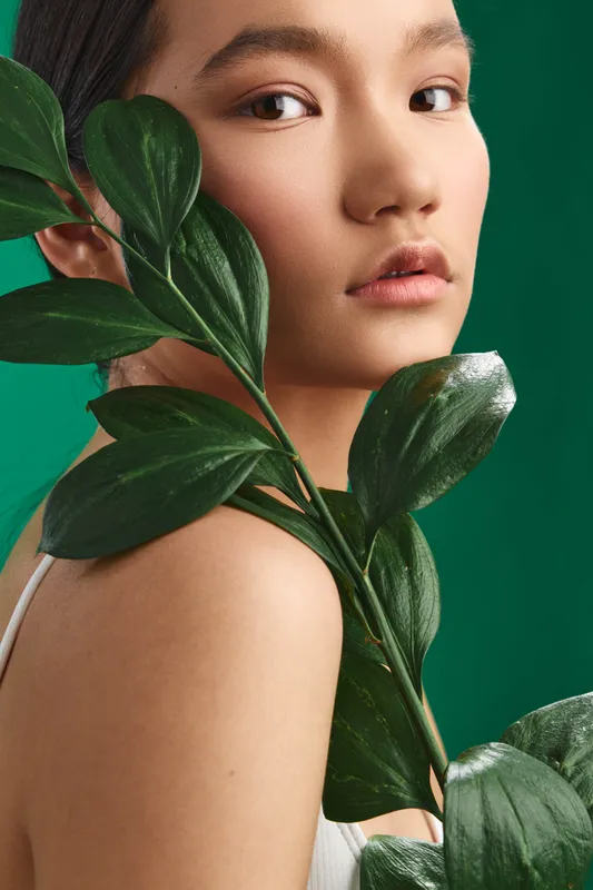 Asian woman models a serene portrait with a leafy stem near her shoulder, soft gaze, and natural glow. The scene conveys calm beauty, fresh style, and subtle botanical elegance.