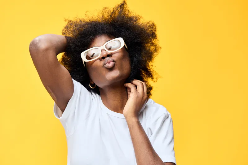 Young Black woman with curly hair playfully posing against a vibrant yellow background, expressing confidence and joy in a casual white t shirt and stylish glasses