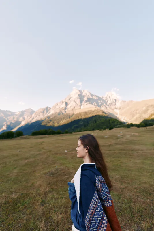 woman profile walking in meadow with mountains in background, landscape view of nature outdoors, young traveler wearing jacket and patterned shawl enjoying peaceful alpine scenery