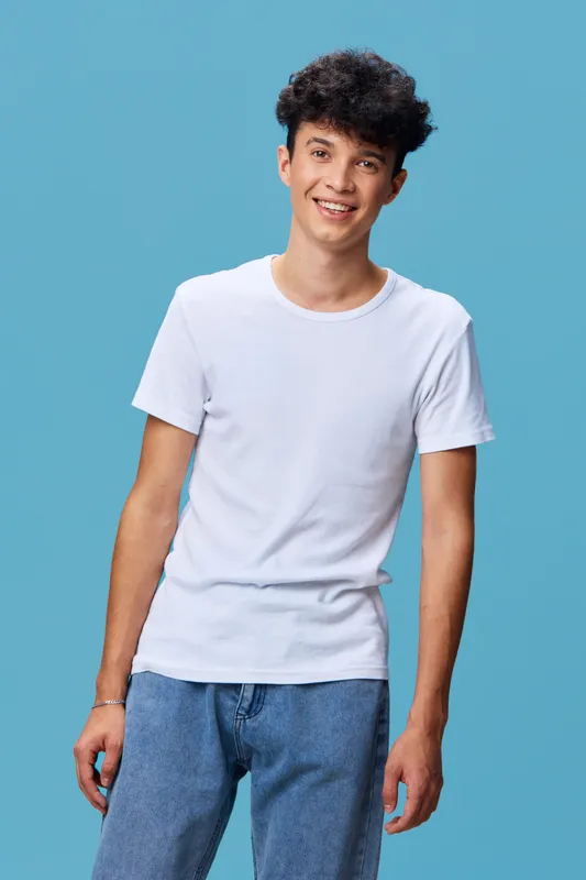 smiling man in white t-shirt and blue jeans stands relaxed against blue background, casual style with friendly expression and curly hair, young adult male portrait