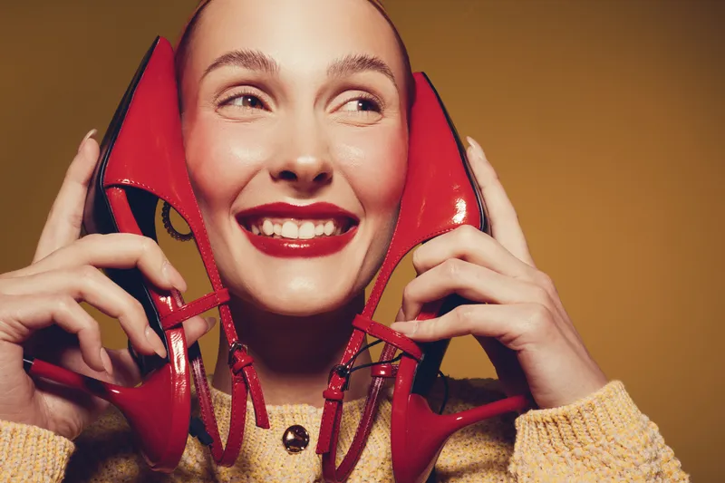 Smiling woman holds red high heel shoes near face, wearing yellow sweater. Close-up portrait with bright makeup and joyful expression on warm brown background. Concept of fashion and happiness.