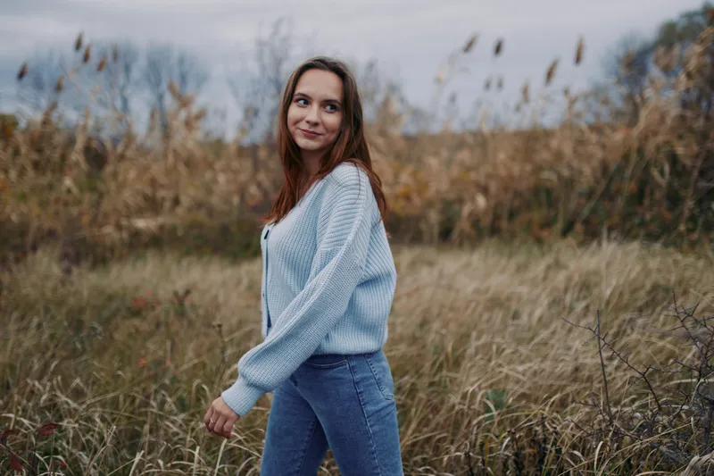 A young woman walking through tall coastal grasses, captured in an authentic, candid moment of simple outdoor life; natural pose, soft light, genuine expression conveying calm and mindful presence.