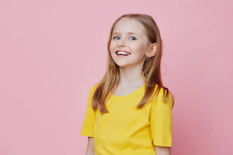 Cheerful young girl with long hair wearing a bright yellow t shirt, smiling against a soft pink background, exemplifying joy and innocence in childhood
