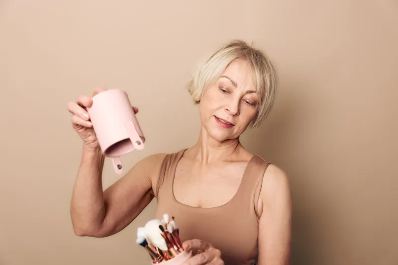 Mature woman with short blonde hair holding a pink container pours makeup brushes in beige studio. Concept of beauty tools, cosmetics storage, and personal care routine for women.