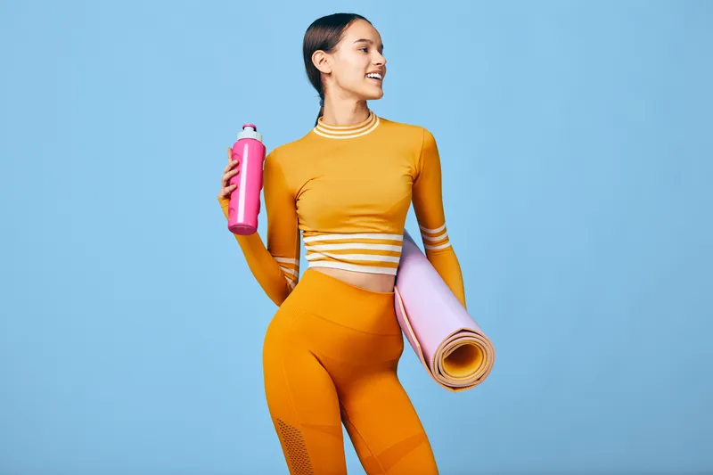 Fit woman in orange activewear holding a pink water bottle and a rolled yoga mat, smiling confidently against a clean blue background, healthy lifestyle and exercise concept image.